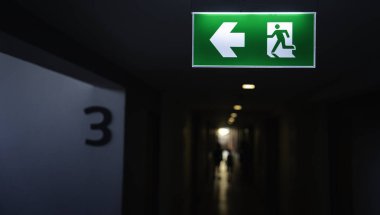Karanlık koridorda koşan adam ikonu ve yön oku olan acil çıkış işareti. Güvenlik tahliye güzergahı yangın çıkış sembolü bina güvenliği ve risk yönetimi kavramı.