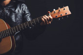muž v kožené bundě hraje na akustickou kytaru na černém pozadí