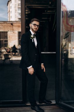 Sakallı, gözlüklü, klasik siyah takım elbiseli, beyaz gömlekli ve papyonlu yakışıklı bir damat. Tam boy erkek portresi. Siyah takım elbiseli şık bir adam.