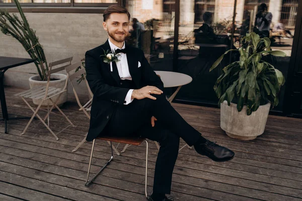 Klasik siyah takım elbiseli, beyaz gömlekli ve papyonlu, yaz terasında sandalyede oturan yakışıklı, mutlu bir damat. Tam boy erkek portresi. Siyah takım elbiseli şık bir adam.