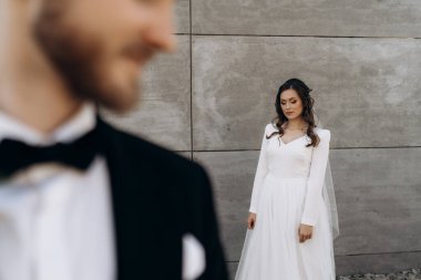 Güzel bir gelin damadın arkasında duruyor. Gelinin tarzı. Güzel gelin klasik beyaz bir elbise ve duvak giyiyor. Klasik siyah takım elbiseli, beyaz gömlekli ve papyonlu damatlık..