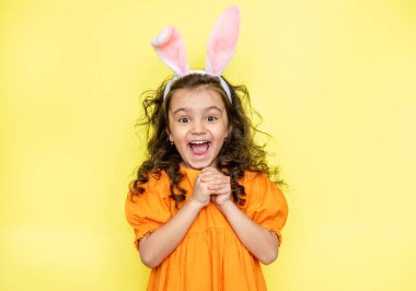 Mutlu kıvırcık kız tavşan kulağı takıyor, tatilin gizemini ve kutlamasını seviyor, turuncu parlak elbiseler giyiyor, sarı arka planda izole edilmiş. Mevsimlik tatil kavramı 