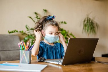 Küçük bir kız, Coronavirus salgını sırasında dizüstü bilgisayarında öğretmenlik yaparken yüz maskesi takıyor.. 