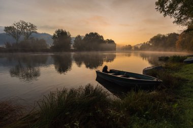 Calm scenic picture of small fishing boat on the Adda river on a hazy sunrise with foliage clipart