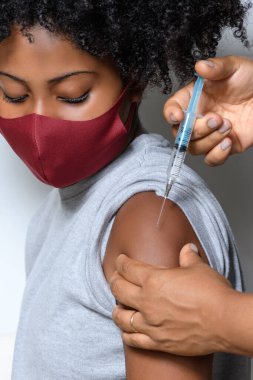 Yüzünde bir gülümseme ile Covid-19 'a karşı koruyucu maske takan siyah bir genç beyaz arka planda izole edilmiş aşı markasını gösteriyor.