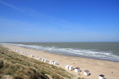 Güneşli bir günde, Belçika 'nın Kuzey Denizi kıyısında geniş kumlu bir sahilde beyaz kulübeler. Bulutsuz bir gökyüzü, yanı başında büyüyen kumullar.