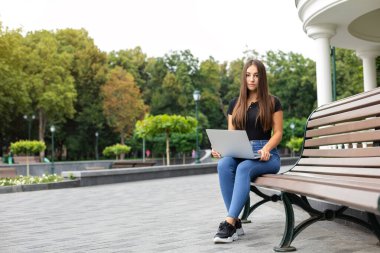 Eğitim, iş dünyası, teknoloji ve internet kavramı - Siyah tişörtlü ve kot pantolonlu gülümseyen Avrupalı kız yazın parkta bankta oturuyor ve elinde dizüstü bilgisayarla.