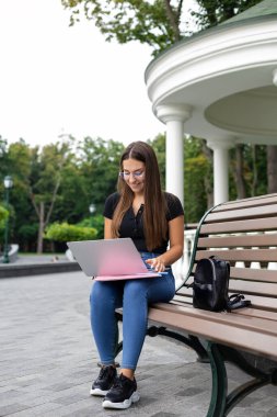Serbest meslek, iş dünyası, teknoloji ve internet konsepti. Yazın parktaki bankta oturan gülümseyen koyu renk saçlı bir kız. Bilgisayarına çanta, not defteri ve kalemle bakıyor..