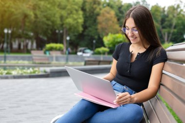 Eğitim, teknoloji ve internet kavramı - Parktaki bankta oturmuş bilgisayarında ders çalışırken sınıf arkadaşlarına bakan koyu renk saçlı gülümseyen bir Avrupalı kız..