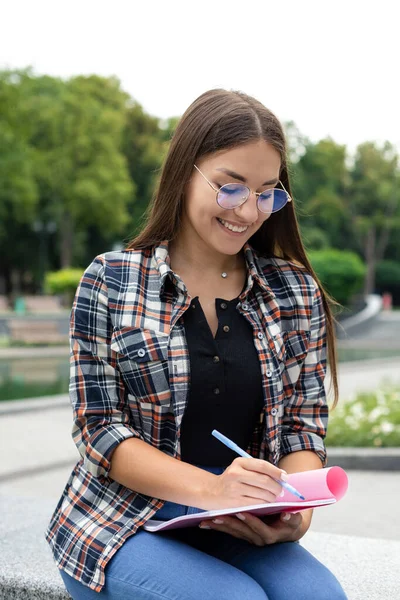 新鮮な空気 再生されたシャツと眼鏡の若い白い女性が公園に座って オンライン会議中に大きなノートブックにメモを書く — ストック写真