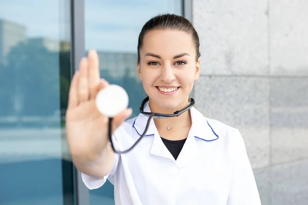 Medicine, healthcare, profession and people concept. European white female doctor in a medical gown shows her stethoscope to the camera and listening something outside in sunny weather