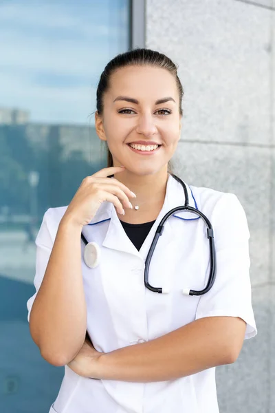 Healthcare, medicine, profession and people concept. Portrait of a happy European white-haired doctor girl with her hand at her face in a medical gown, wearing a stethoscope outside in sunny weather