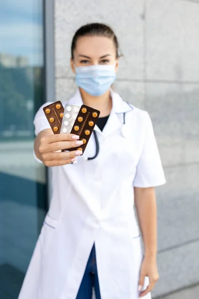 Medicine, healthcare, profession and people concept. A European girl doctor in a white coat with a stethoscope and a virus mask, showing to the camera several packages of pills in her hands outside