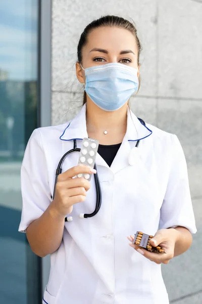 Medicine, healthcare, profession and people concept. A European girl doctor in a white coat with a stethoscope and a virus mask holding several packs of pills