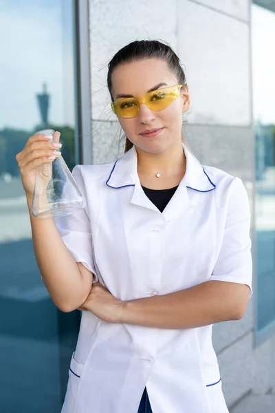 Sciene, chemistry, biology and medicine concept. Oriental dark-haired female chemist in a white coat and yellow glasses holding an Erlenmeyer flask outside and looking at the camera.