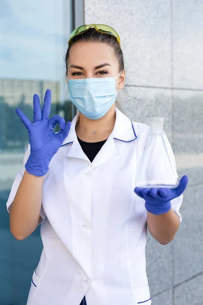 Sciene, chemistry, biology and medicine concept. Oriental dark-haired female scientist in medical gown, protective mask and rubber gloves holding Erlenmeyer flask and showing ok hand sign outside.