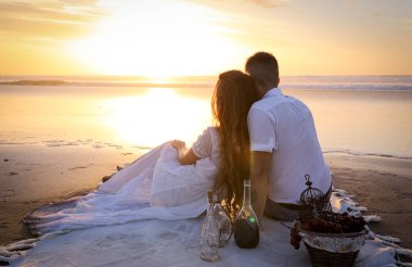Sihirli günbatımında okyanusta romantik bir randevu. Beyaz elbiseli bir kız ve erkek sihirli bir gün batımında okyanusta beyaz bir yatak örtüsüne oturur ve güzel bardaklardan su içerler..