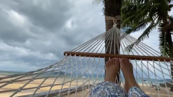 Frauenfüße schwingen in einer Hängematte und entspannen sich unter Kokospalmen am Strand. — Stockvideo