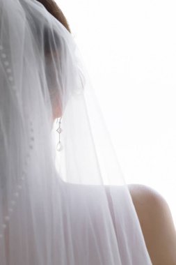 Gelinin silueti arkadan, küpeli ve duvağı beyaz arka planda.