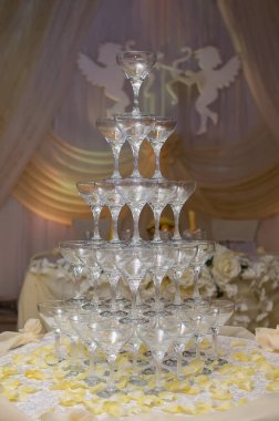 Şampanya kadehleri piramidi gül yapraklarıyla süslenmiş bir masanın üzerinde duruyor. Arka planda melekler var.