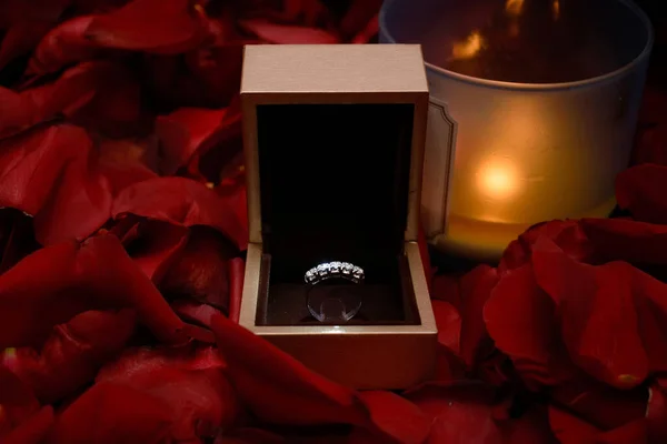 Silver ring in a wooden box, red rose petals around it, a glass with a burning candle next to it