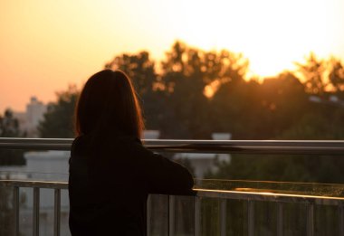 Kız ağaçların arasından gün batımını seyrediyor, cam bir köprüde duruyor, arkadan dikiz aynasından.