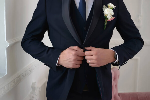 Groom in a dark blue suit with a tie and a boutonniere fastens a button on his suit close-up