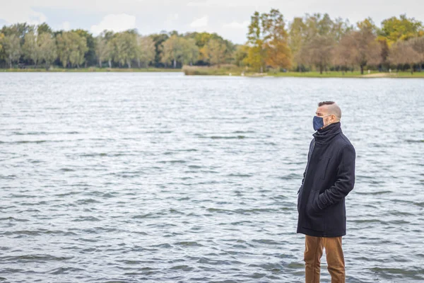 Maskeli bir adam gölün kenarındaki parkta duruyor.