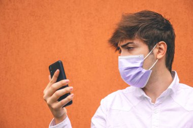 Mor maskeli adam turuncu arka planda akıllı telefonuna bakıyor..