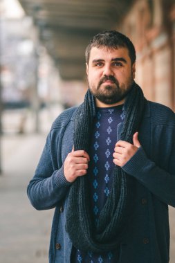 Mavi kazaklı sakallı adam, elleri eşarbında caddede duruyor, kameraya bakıyor.. 