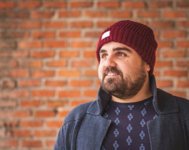 Tuğla duvarda gülümseyen yün şapkalı ve kazaklı bir adam..