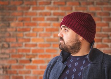 Yünlü şapkalı ve kazaklı bir adam tuğla duvara bakıyor..