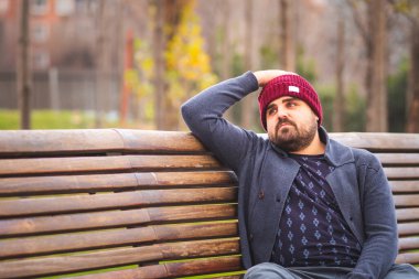 Yünlü şapkası ve kazağı olan, parktaki bankta oturmuş endişeli bir adam..