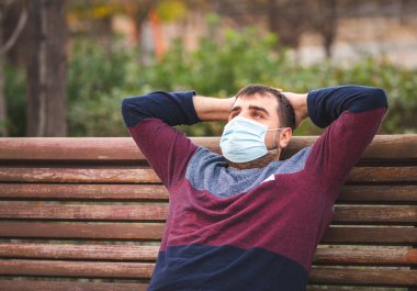 Koruyucu maskeli adam gün batımında bankta oturmuş dinleniyor.. 