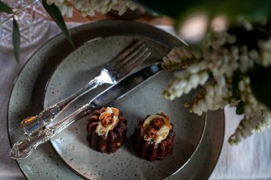 Çatal ve bıçaklı seramik tabakta çikolatalı çörekler, ahşap masada tekstil peçetesi ve çiçekler.