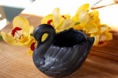 Siyah kuğu şeklinde seramik vazo, ahşap bir masada sarı orkideler.