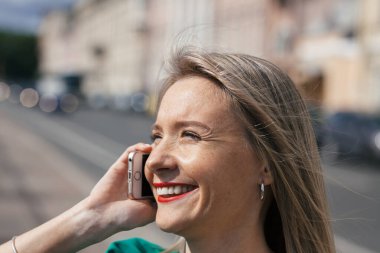 Yakından çekilmiş bir portre. Kırmızı rujlu, yeşil elbiseli, güzel, sarışın bir kadın. Nehrin kenarında, cep telefonuyla konuşuyor.