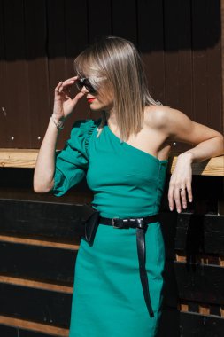 Şık yeşil elbiseli, kırmızı rujlu ve güneş gözlüklü şık, genç, şık sarışın kadın şehir ortamında poz veriyor.