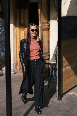 Genç, güzel, güler yüzlü, sarışın, güneş gözlüklü, siyah pantolonlu, deri ceketli, şehir ortamında binaların önünde poz veren bir kadın.