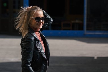 Genç, güzel, güler yüzlü, sarışın, güneş gözlüklü, siyah pantolonlu, deri ceketli, şehir ortamında binaların önünde poz veren bir kadın.