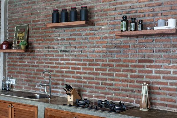 spacious kitchen with red brick wall, gas stove, sink and jars on the shelves