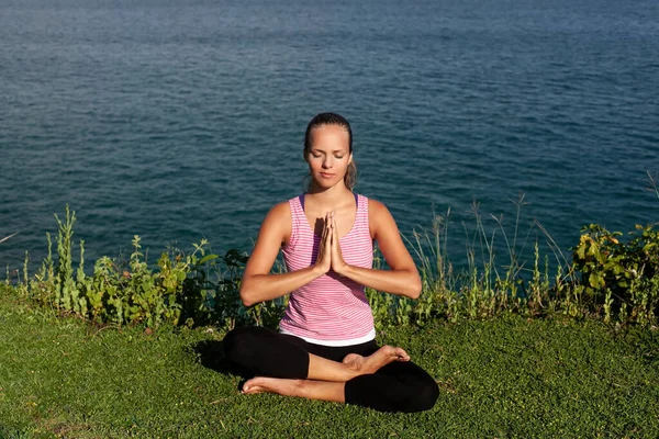 Spor kıyafetli güzel, gülümseyen sarışın kadın arka planda mavi denizle çimlerin üzerinde yoga yapıyor.