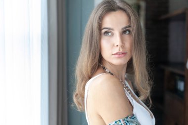 Pencerenin yanındaki odada uzun saçlı, beyaz bluzlu güzel, genç esmer kadın.