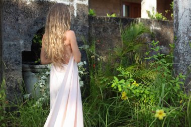 Eski binanın duvarının yanında yeşil ve çiçekli, uzun dalgalı sarı saçlı, beyaz uzun elbiseli genç bir kadının arka manzarası.