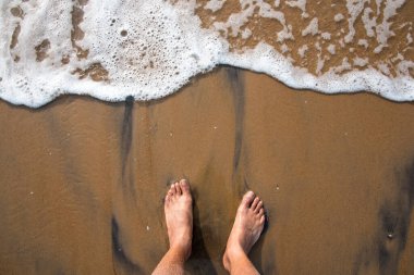 Dalgalar yaklaşırken deniz kıyısındaki ıslak kumda ayakların üst görüntüsü..