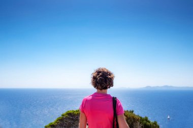 Sırtüstü uzanmış pembe tişörtlü genç bir kadın arka planda deniz olan bir uçurumdan ufku inceliyor.