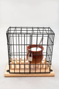 clay jar full of sugar with a teaspoon inside, locked in a prison with a lock on the door, concept of forbidden, diet or weight loss, vertical
