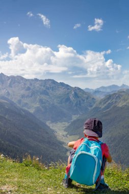 Sırt üstü çömelmiş mavi şapkalı ve sırt çantalı maceraperest ve korkusuz küçük çocuk dağın tepesinden bir dağ manzarası vadisini dikey olarak gözlemler.