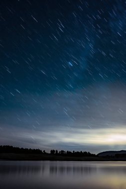 Gece manzarası yıldızlı bir gökyüzü, arka planda bir ormanın silueti ve ön planda dikey bir göl.