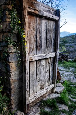 Eski ahşap kapı bir tarafı küçük sarı çiçekli köy evinden kısmen açılmış ve arka planda kırsal manzarası var.